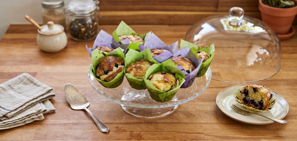 Ein Arrangement von frisch gebackenen Blaubeer-Muffins mit Streusel, die in bunten Papierformen auf einer gläsernen Etagere auf einer Holztheke angerichtet sind, umgeben von Küchenutensilien und einer Glasglocke.