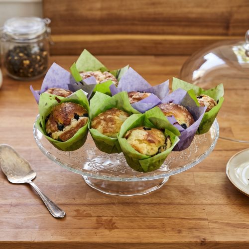 Ein Arrangement von frisch gebackenen Blaubeer-Muffins mit Streusel, die in bunten Papierformen auf einer gläsernen Etagere auf einer Holztheke angerichtet sind, umgeben von Küchenutensilien und einer Glasglocke.
