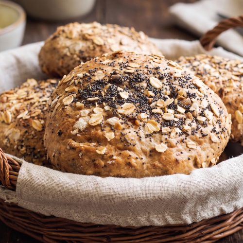 Goldbraun ausgebackene Haferflockenbrötchen mit Mohn und Haferflocken-Topping, serviert in einem geflochtenen Brotkorb mit Leinentuch.
