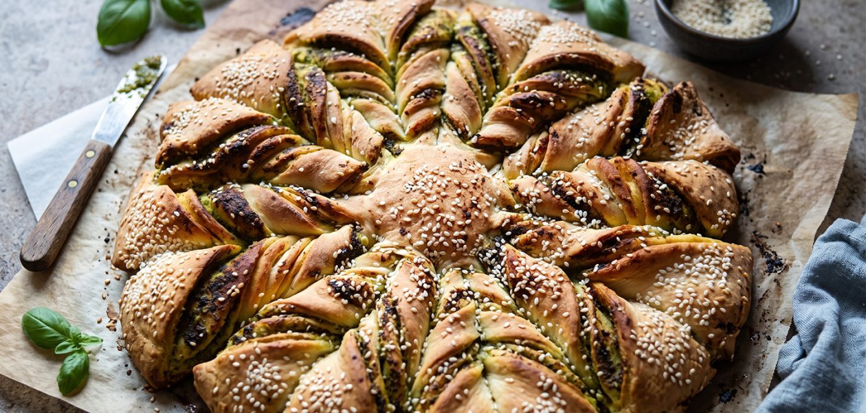 Frisch gebackener, goldbrauner Brotstern mit grünem Pesto und Sesam auf Backpapier, serviert mit frischem Basilikum.